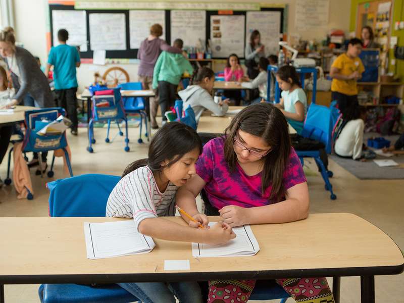 Two girls collaborate on assignments at a classroom table, while other students engage in various activities in the background.