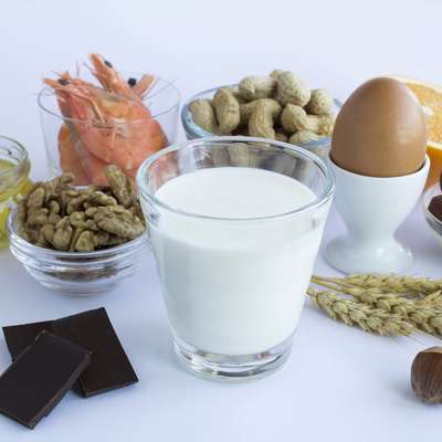 A variety of foods including shrimp, nuts, an egg, milk, chocolate, and an orange are arranged on a light background.