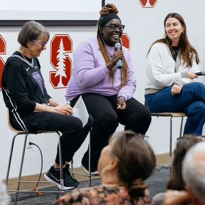 Four women participate in a panel discussion, seated on stools with microphones, sharing experiences and insights in a community setting.