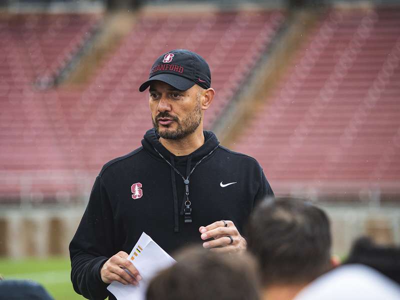 Stanford coach in a black cap and hoodie, holding papers in one hand, speaks to a group of players in an empty stadium.