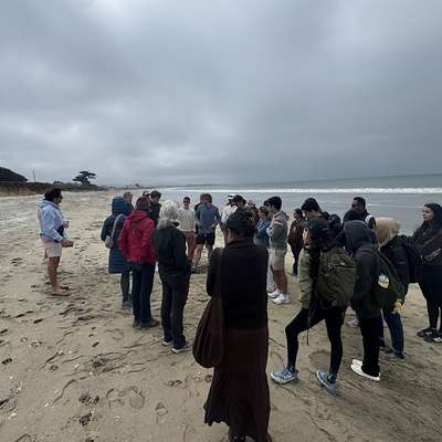 Students and instructors gather on a beach in Half Moon Bay under gray, overcast skies, listening to a speaker.
