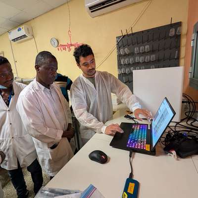 Group of people in lab coats observe a technician using a laptop in a laboratory setting.