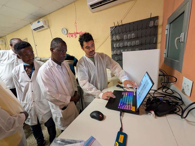 Group of people in lab coats observe a technician using a laptop in a laboratory setting.