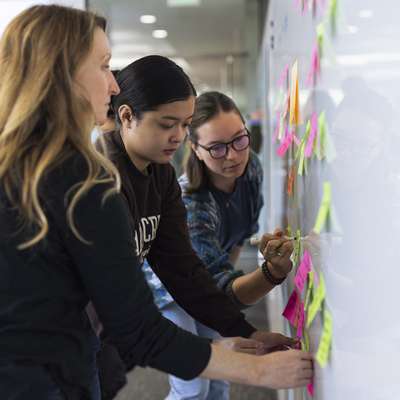 Three people are writing on a whiteboard. The whiteboard also has many colorful sticky notes on it.