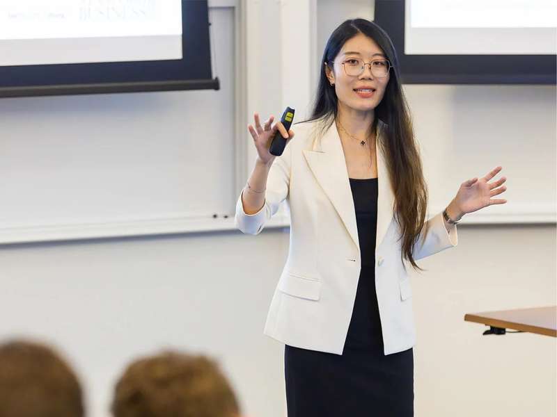 Yuyan Wang presents to an audience, holding a remote in one hand, in a classroom setting.