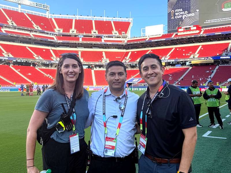 Alyssa Perry, Shashank Ravi, and Eli Carrillo smile together on the field of a stadium with empty red seats and World Cup signage.