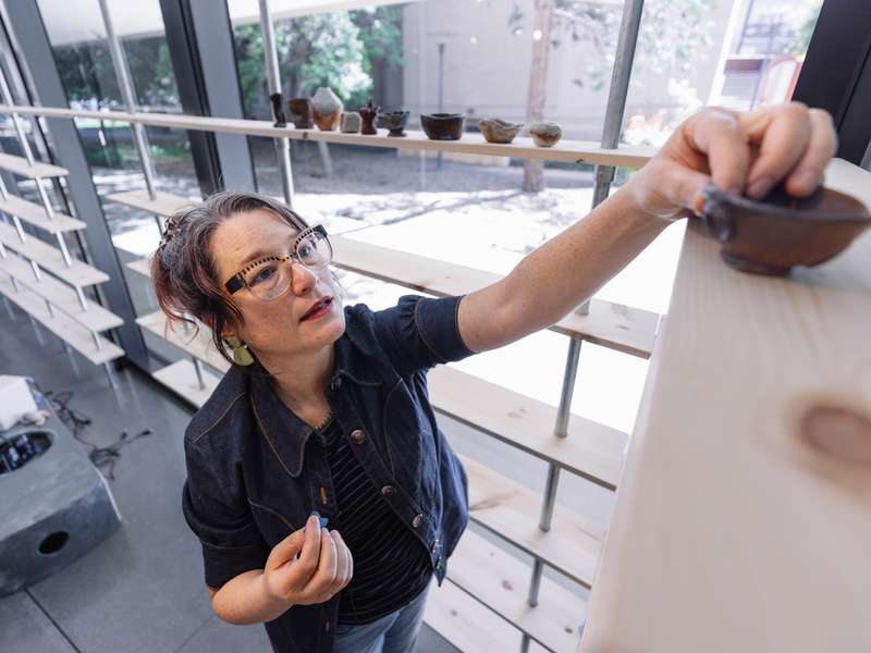 Artist places a bowl on a high shelf in a bright studio with pottery displayed on shelves in the background.