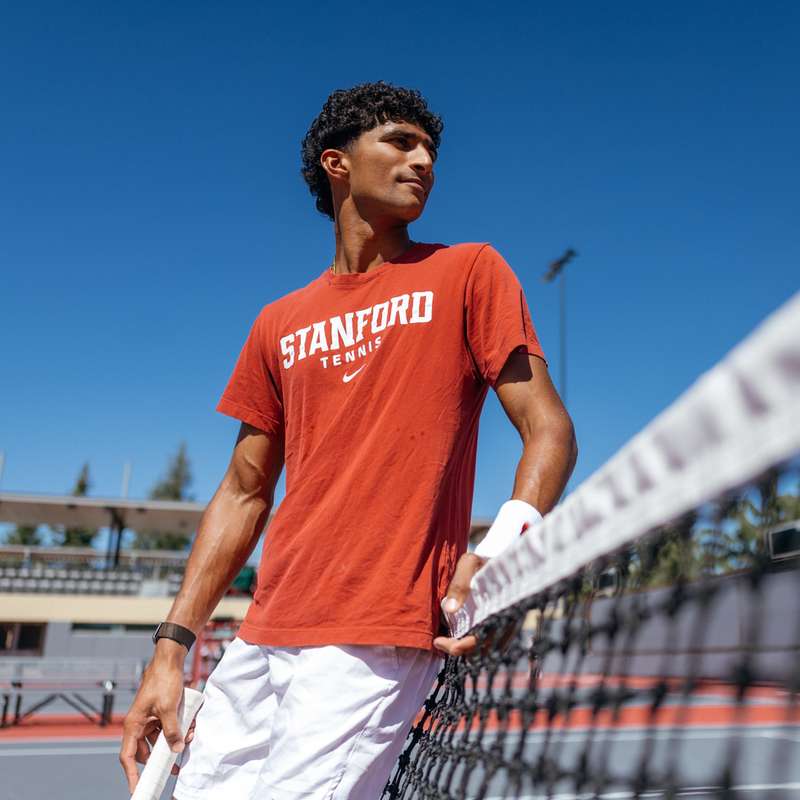 Samir Banjeree in a Stanford tennis shirt leans against the net on a tennis court under clear blue skies.