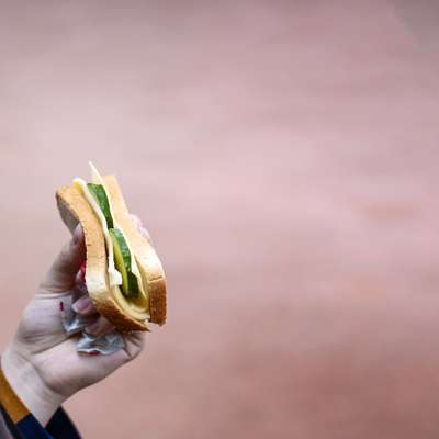 A person holds a sandwich with cheese and vegetables, against a blurred background.