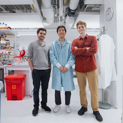 Three researchers stand together smiling in a bright, well-stocked lab, one wearing a blue lab coat.