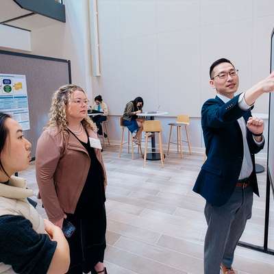 A presenter discusses a research poster while two audience members listen attentively in a conference setting.