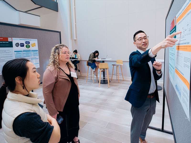 A presenter discusses a research poster while two audience members listen attentively in a conference setting.