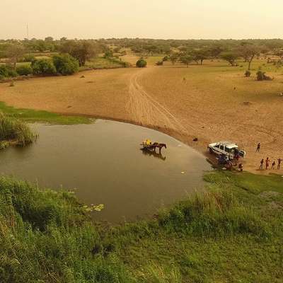 Aerial view of a riverbank with a horse and cart in shallow water, and people gathered along the shore near a parked vehicle.