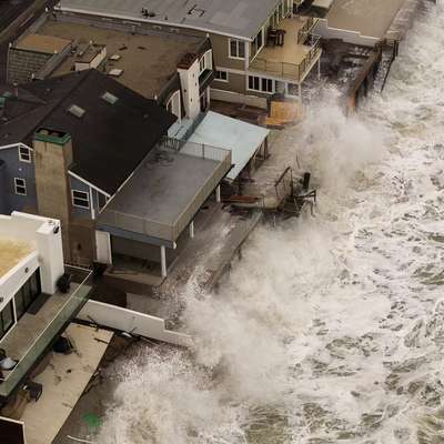 Aerial view of large waves battering oceanfront homes, with storm damage visible on decks and walkways.