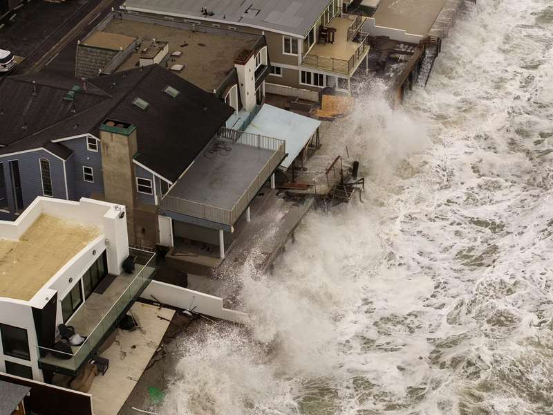Aerial view of large waves battering oceanfront homes, with storm damage visible on decks and walkways.