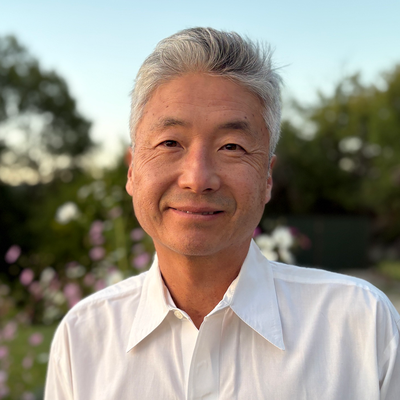 Man with gray hair in a white shirt smiling outdoors with trees and flowers in the background.