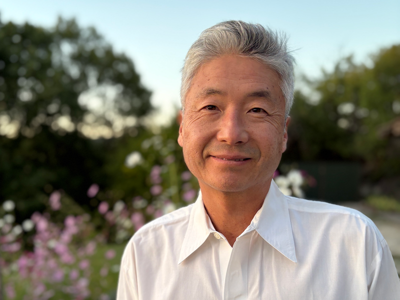 Man with gray hair in a white shirt smiling outdoors with trees and flowers in the background.