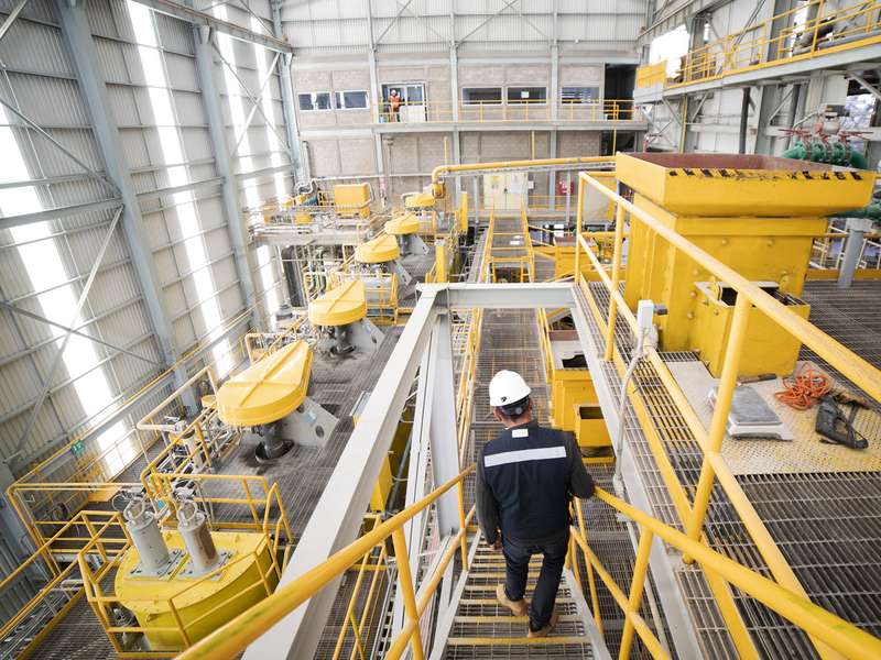 Worker in a hard hat walks down stairs past yellow flotation cells and catwalks inside a large mineral processing plant.