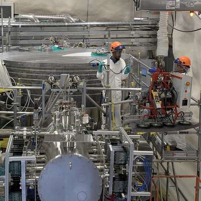 Two workers in white coveralls and orange hard hats work on scaffolding beside large stainless steel tanks and piping.