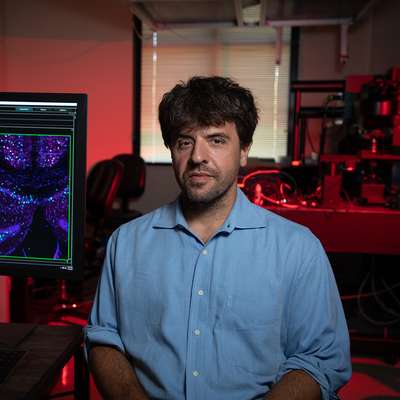 Karl Deisseroth sits in a red-lit lab beside a monitor showing a purple and teal microscope image.