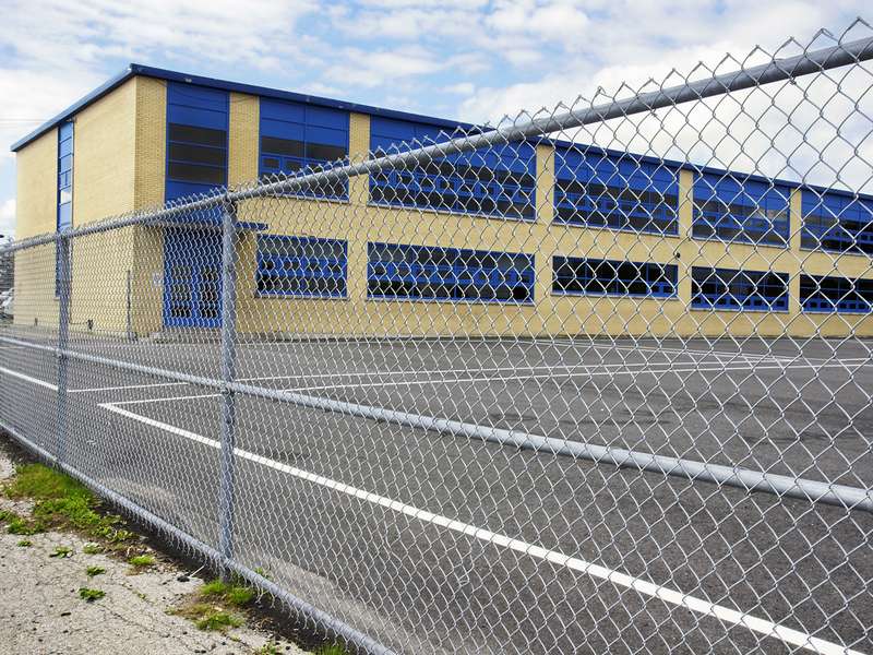 A school building with blue windows is seen behind a chain-link fence.