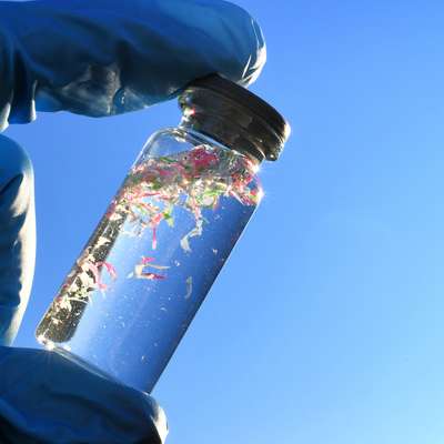 A gloved hand holds a small glass vial of clear liquid with pink, green and white plastic fragments against blue sky.