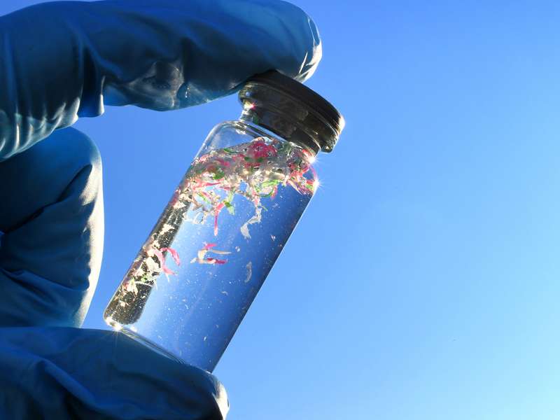 A gloved hand holds a small glass vial of clear liquid with pink, green and white plastic fragments against blue sky.