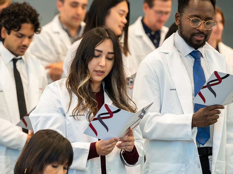 Graduate students in white coats stand in rows holding printed programs with a DNA helix design, reading aloud at a coat ceremony.