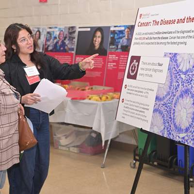 A staff member wearing a name badge explains a cancer research poster's key statistics to a visitor at a health fair.