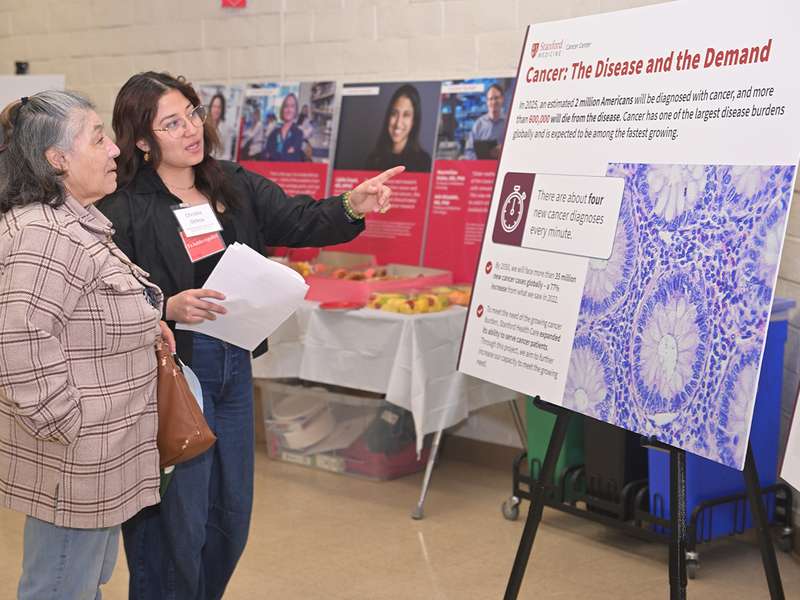 A staff member wearing a name badge explains a cancer research poster's key statistics to a visitor at a health fair.