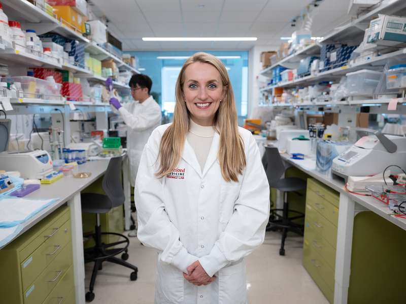 Agnieszka Czechowicz in a lab coat standing in a laboratory filled with equipment and materials, while a colleague works in the background.