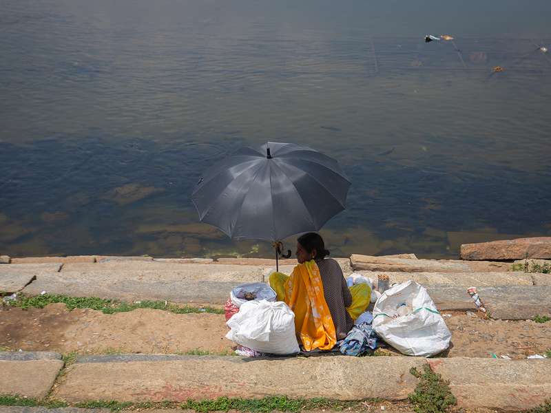 A woman sits by a riverbank under a black umbrella, surrounded by bags.
