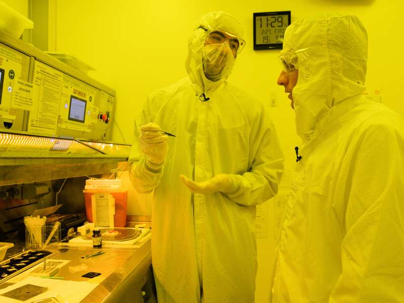 Two people in protective gear discuss procedures near specialized lab equipment, with various tools and documents visible on the table.