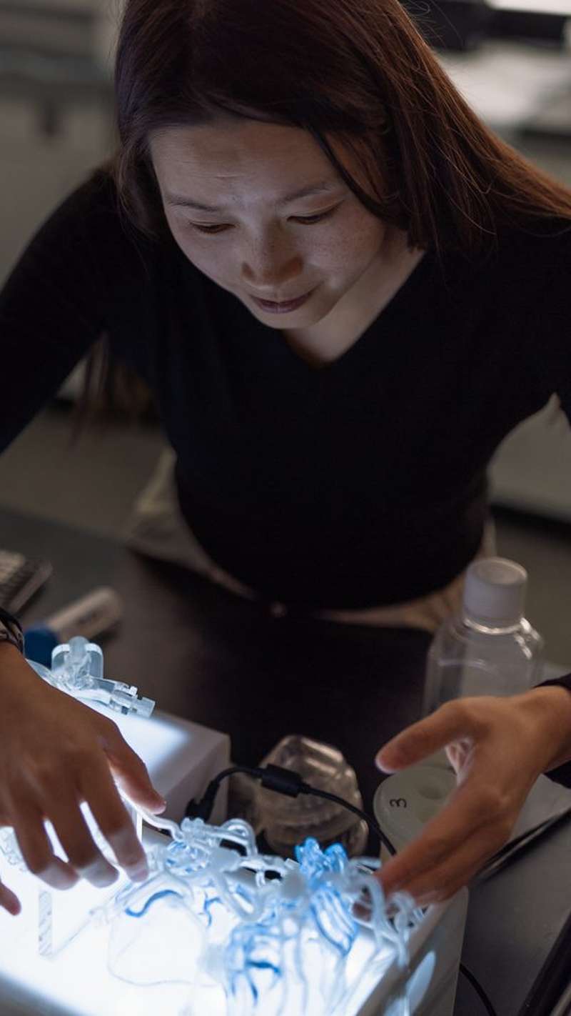 Researcher Renee Zhao leans over a glowing light table in a dark lab, examining a tangle of clear and blue 3D-printed tubing with both hands.