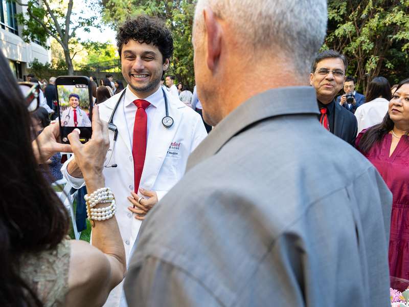A medical student in a white coat smiles as a family member takes his photo at an outdoor celebration.