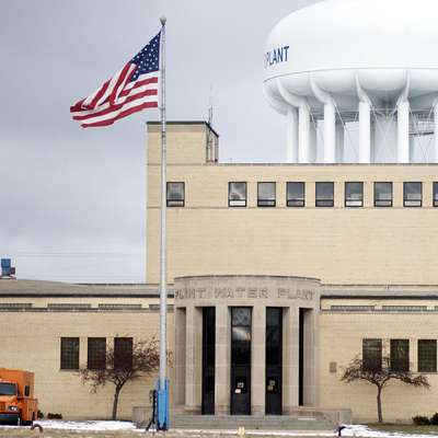 The Flint Water Plant, a tan brick building with a large white water tower behind it and a U.S. flag flying out front under an overcast sky.