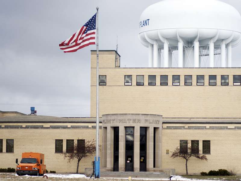 The Flint Water Plant, a tan brick building with a large white water tower behind it and a U.S. flag flying out front under an overcast sky.