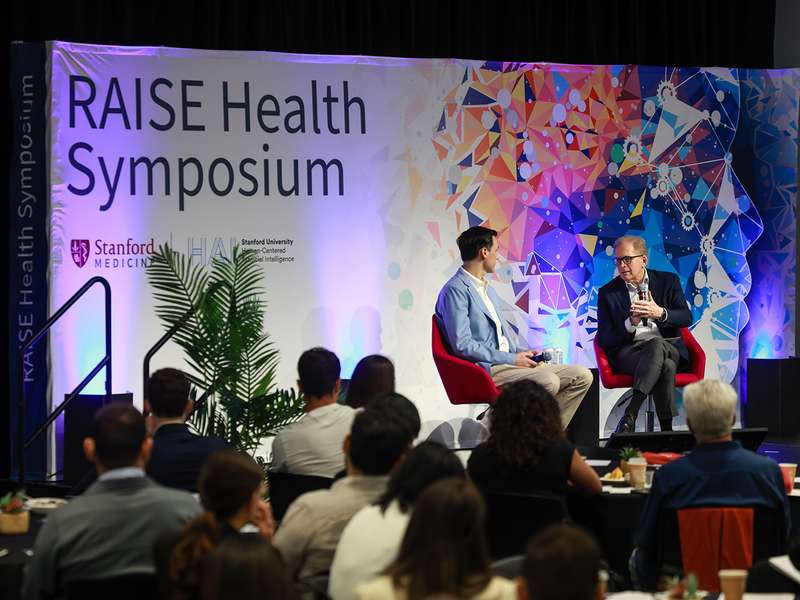 Two speakers discuss topics on stage at the RAISE Health Symposium.