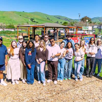 Students and staff standing together in a community garden with green hills in the background.
