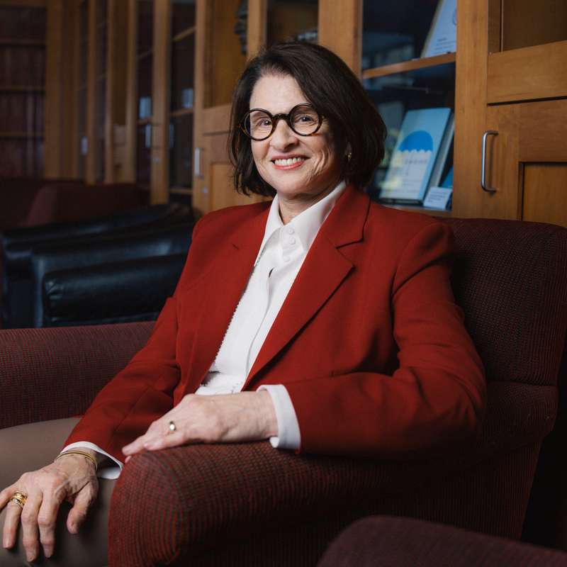 Laura Carstensen in a red blazer and white blouse sits in an armchair, smiling for the camera, against wood-paneled bookshelves in the background.