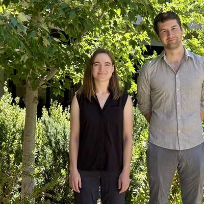 Melissa Gray in a black sleeveless top and Steven Banik in a gray button-down shirt stand outdoors among sunlit trees and shrubs.