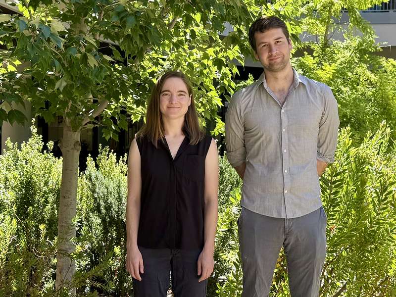 Melissa Gray in a black sleeveless top and Steven Banik in a gray button-down shirt stand outdoors among sunlit trees and shrubs.
