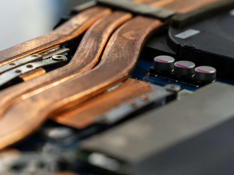 Macro shot of copper heat pipes on a laptop's circuit board, with capacitors and screws visible, shallow depth of field.