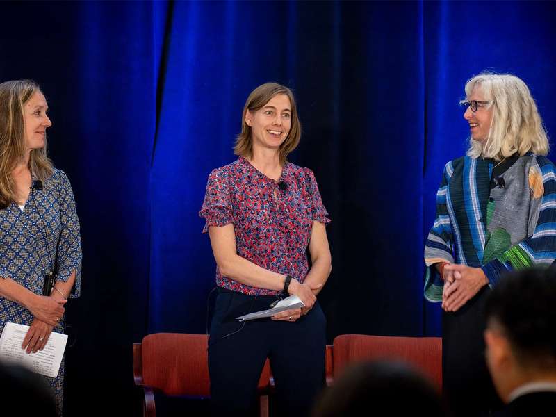 Three women engage in discussion on stage at an event, with microphones and seated chairs visible in the background.