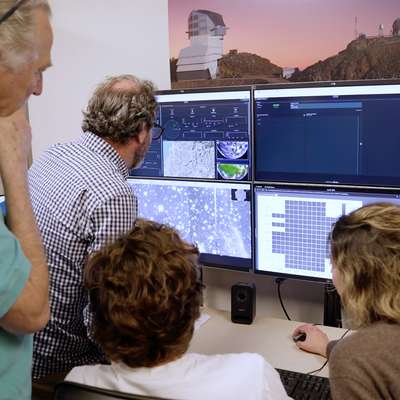 A group of four people studies data on multiple computer screens in an office setting, with an observatory visible in the background.