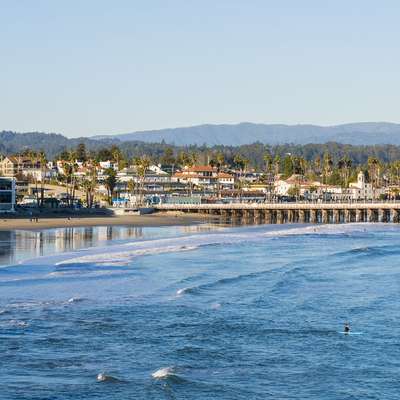 Coastal scene of Santa Cruz, CA, featuring a beach, pier, and palm trees under a blue sky, with a surfer in the water.