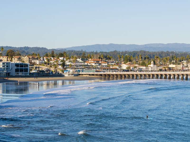 Coastal scene of Santa Cruz, CA, featuring a beach, pier, and palm trees under a blue sky, with a surfer in the water.