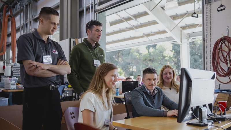 A group of students collaborates and observes while working on a computer in an educational workshop setting.