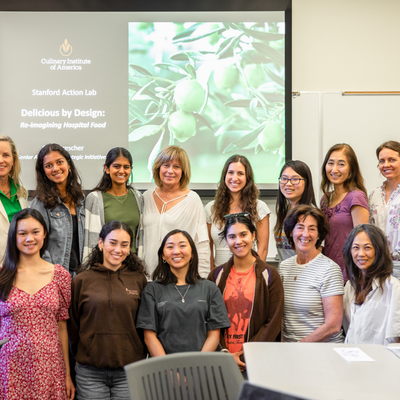 Group of students and faculty pose in a classroom before a slide reading "Delicious by Design: Re-Imagining Hospital Food."