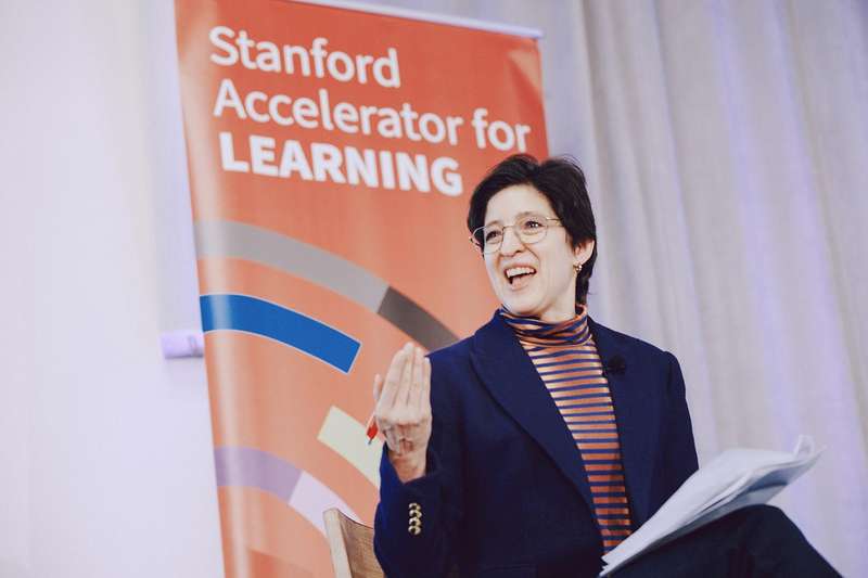 A woman gesticulates while speaking to an audience, in front of a sign that says "Stanford Accelerator for Learning"